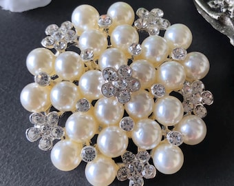 May include: A cream-coloured brooch with a cluster of faux pearls and sparkling rhinestone accents. The brooch features a floral design with multiple pearl beads and small, silver-toned flower-shaped embellishments.
