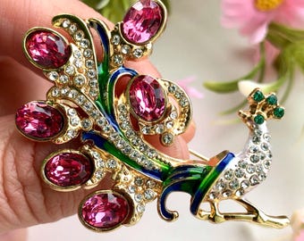 May include: A gold peacock brooch featuring pink oval gemstones and clear crystals. The peacock's tail feathers are detailed with green and blue enamel. The bird's body is white with crystal accents.