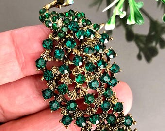 May include: A peacock brooch with a gold-tone metal frame and emerald green rhinestones. The brooch features a detailed design with a fan-shaped tail and a curved neck. The peacock's head is adorned with iridescent rhinestones.