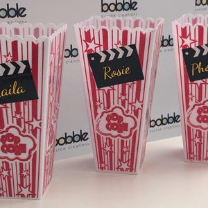 May include: Three red and white popcorn containers with black and white clapperboard tags. The tags have the names "Laila", "Rosie", and "Phoebie" written on them.