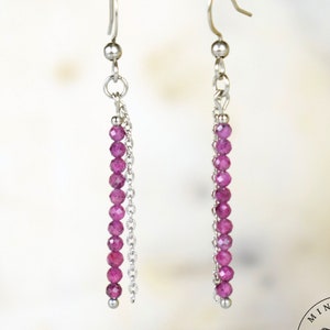 May include: A pair of dangle earrings with a silver-tone metal design. Each earring features a chain and a strand of small, round, magenta-coloured beads. The earrings are suspended from a silver-tone hook.