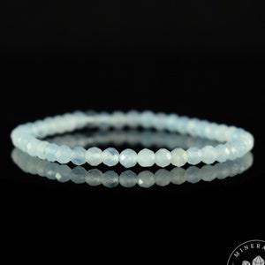 May include: A delicate aquamarine beaded bracelet. The light blue gemstones are faceted and strung together, creating a subtle sparkle. The bracelet is displayed on a reflective black surface, highlighting its color and design.