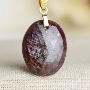 Op de afbeelding: Een ovale hangerketting met een gefacetteerde, diep bordeauxrode edelsteen en gouden spikkels. De edelsteen hangt aan een fijne gouden ketting. De hanger heeft een gouden balein. De achtergrond is neutraal.