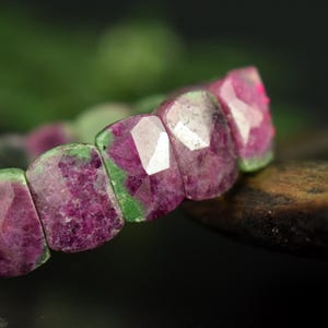 Op de afbeelding: Close-up van een armband met gefacetteerde robijn-zoisiet kralen. De kralen hebben een mix van diepe roze en groene tinten, met een geometrische snit die het licht vangt. De armband is geplaatst tegen een vage groene en zwarte achtergrond.