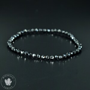 May include: A black beaded bracelet with faceted beads. The bracelet is made of small, dark gray beads strung together. The beads have a reflective quality, catching the light. The bracelet is displayed on a dark surface. The logo "MINERAUX VERTUS" is in the lower left corner.