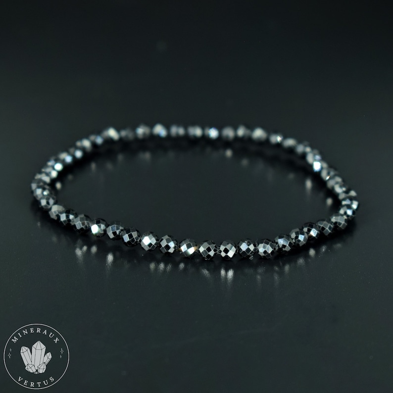 May include: A black beaded bracelet with faceted beads. The bracelet is made of small, dark gray beads strung together. The beads have a reflective quality, catching the light. The bracelet is displayed on a dark surface. The logo "MINERAUX VERTUS" is in the lower left corner.