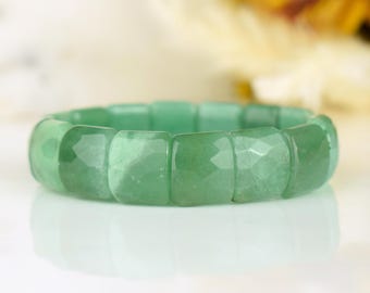 Può includere: Un braccialetto di avventurina verde. Il braccialetto è composto da perline quadrate sfaccettate. Le perline sono di un colore verde chiaro traslucido. Il braccialetto è esposto su una superficie bianca con uno sfondo morbido.