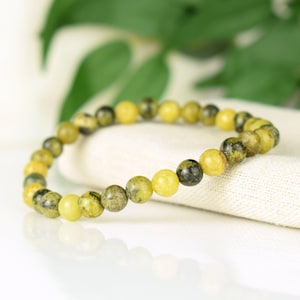May include: A beaded bracelet featuring a series of round, yellow and olive-green beads. The bracelet is resting on a white surface, with green foliage in the background. The beads have a diameter of approximately 0.3 inches.