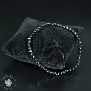 May include: A black beaded bracelet with faceted beads is displayed on a black velvet pillow. The bracelet is circular and appears to be made of small, evenly sized beads. The background is black, creating a high-contrast image.