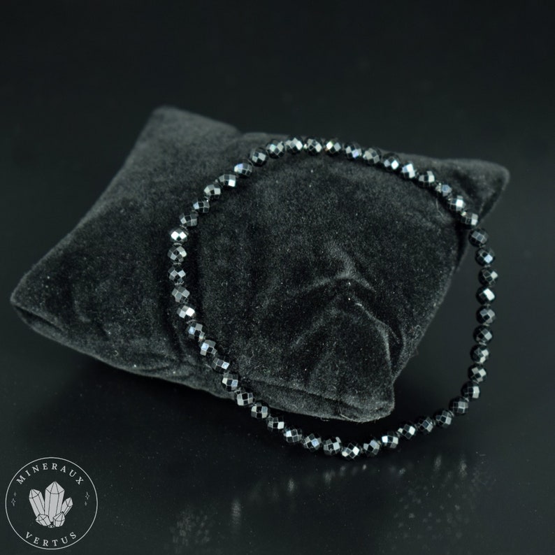May include: A black beaded bracelet with faceted beads is displayed on a black velvet pillow. The bracelet is circular and appears to be made of small, evenly sized beads. The background is black, creating a high-contrast image.