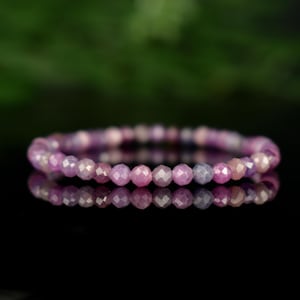 May include: A beaded bracelet featuring faceted, round gemstones in shades of pink and purple. The bracelet is displayed on a reflective black surface, with a blurred green background. The gemstones appear to be strung together on an elastic band.
