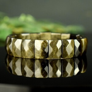 May include: A stretch bracelet composed of rectangular, faceted beads with a metallic gold and bronze sheen. The beads feature a geometric, multi-faceted surface that reflects light, creating a textured appearance across the entire circular band.