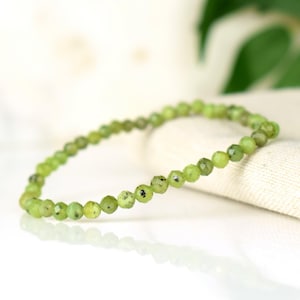 May include: A delicate bracelet featuring small, faceted, round beads in shades of light green. The beads have subtle dark speckles, and the bracelet is strung on an elastic band. The jewelry is displayed on a white surface.