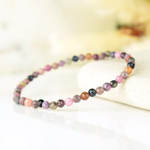 Peut inclure: Un bracelet délicat composé de petites perles facettées de différentes couleurs, notamment rose, violet, marron et bleu. Les perles sont enfilées pour former un bracelet souple. Le bracelet est présenté sur une surface blanche.