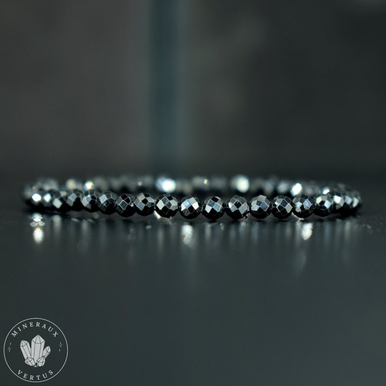 May include: A black beaded bracelet with faceted beads. The bracelet is made of small, round, dark beads strung together. The beads have a reflective, shiny surface. The bracelet is displayed on a dark surface.