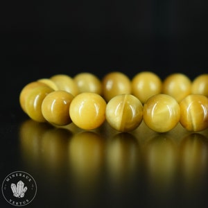 Può includere: Un braccialetto di perline sferiche giallo dorato. Le perline hanno un sottile motivo a strisce e sono molto lucide. Il braccialetto è esposto su uno sfondo nero. Il logo "MINERAUX VERTUS" è visibile.