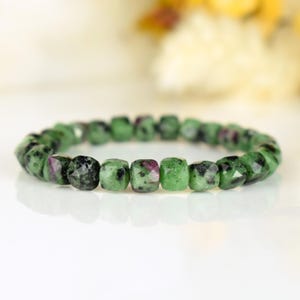 May include: A bracelet made of square-cut gemstones. The bracelet features green and black stones with flashes of red. The gemstones are strung together in a circular design. The bracelet is presented on a white surface.
