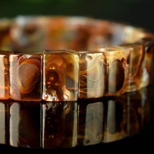 Puede incluir: Primer plano de una pulsera hecha de cuentas rectangulares translúcidas. Las cuentas tienen un aspecto veteado con tonos marrones, ámbar y crema. La pulsera se muestra sobre una superficie reflectante.