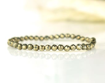 May include: A pyrite beaded bracelet. The bracelet features small, faceted, golden-coloured beads strung together. The beads have a metallic sheen and are arranged in a circular shape. The bracelet is displayed on a white surface.