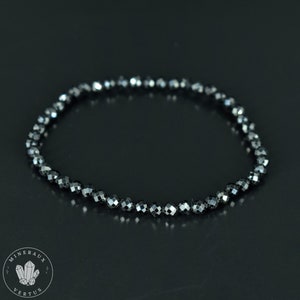 May include: A black beaded bracelet with faceted beads. The bracelet is made of small, dark gray beads strung together, creating a flexible, circular shape. The beads reflect light, giving them a subtle sparkle. The bracelet is displayed on a dark surface.