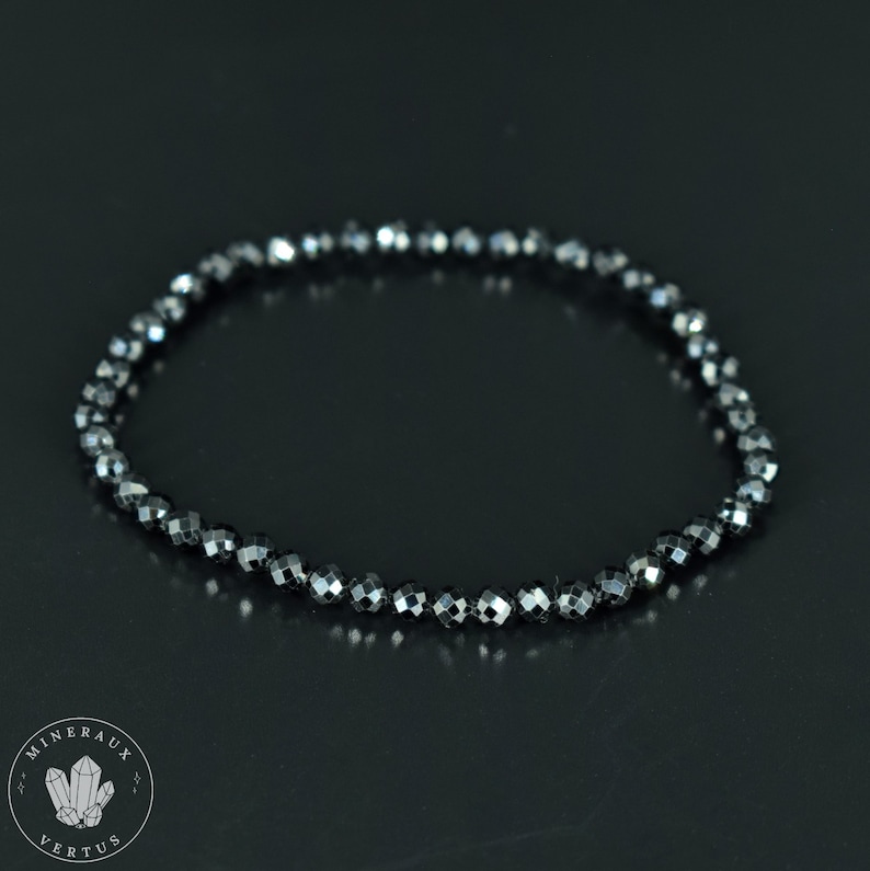 May include: A black beaded bracelet with faceted beads. The bracelet is made of small, dark gray beads strung together, creating a flexible, circular shape. The beads reflect light, giving them a subtle sparkle. The bracelet is displayed on a dark surface.