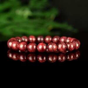 Peut inclure: Un bracelet de perles rondes, rouge foncé et mouchetées. Les perles sont enfilées pour former un cercle complet. Le bracelet est présenté sur une surface noire réfléchissante, avec un fond vert.