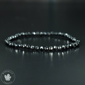 May include: A dark gray beaded bracelet with faceted beads. The bracelet is made of small, round beads strung together, creating a flexible band. The beads have a reflective, metallic sheen, and the bracelet is displayed on a dark surface.