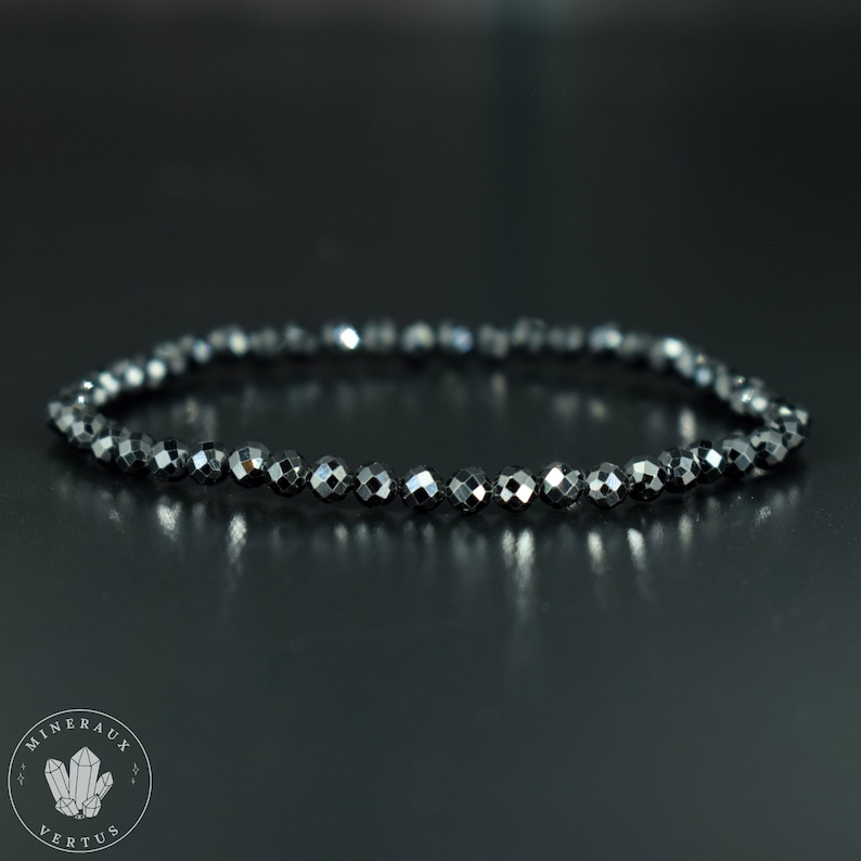 May include: A dark gray beaded bracelet with faceted beads. The bracelet is made of small, round beads strung together, creating a flexible band. The beads have a reflective, metallic sheen, and the bracelet is displayed on a dark surface.