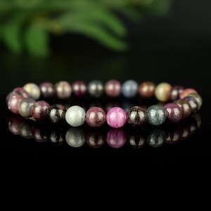 May include: A stretch bracelet composed of round, polished gemstone beads in a variety of colors including shades of pink, deep red, gray, blue, and cream. The beads are arranged in a circular pattern, reflecting on a dark, glossy surface.