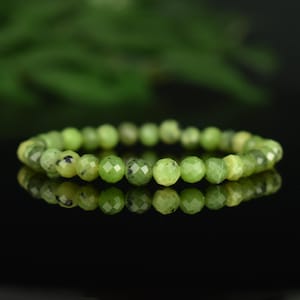 May include: A beaded bracelet featuring faceted, round, light green beads with dark green speckles. The bracelet is displayed on a reflective black surface, with a blurred green background.