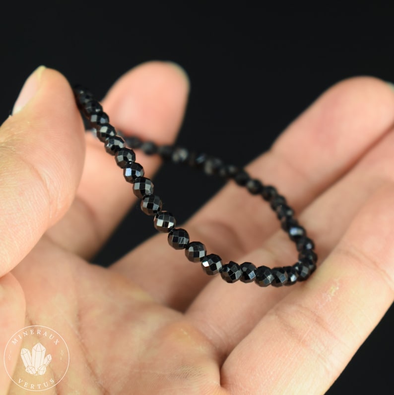 May include: A delicate black beaded bracelet with faceted beads. The bracelet is held in a hand against a black background. The beads are small and evenly sized, creating a sleek and elegant look. The bracelet appears to be strung on a thin, flexible cord.