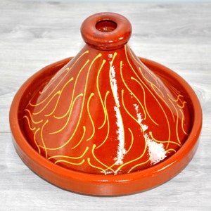 Puede incluir: Una tagine de barro rojo con un diseño de espiral amarillo. La tagine tiene una tapa cónica y una base redonda.