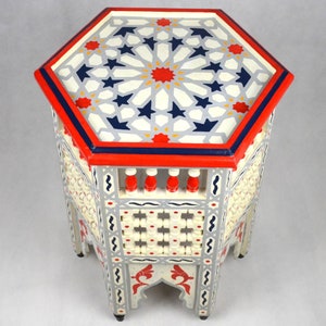 May include: A hexagonal wooden table with intricate geometric designs painted in white, red, and blue. The table has a latticework base and decorative legs.