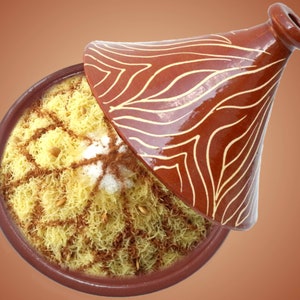 Può includere: Una tagine marocchina tradizionale con una pentola di terracotta marrone e un coperchio con un motivo bianco e marrone. La pentola è piena di un piatto di noodles gialli ricoperti di cannella e pinoli.