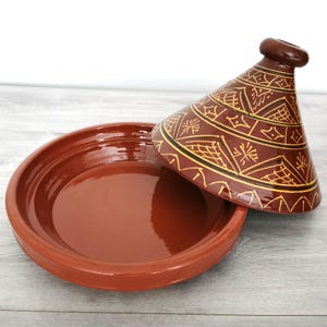 Può includere: Una tagine in terracotta con una finitura smaltata marrone e intricati motivi geometrici in oro. Il coperchio è a forma di cono e ha un piccolo pomo in cima.