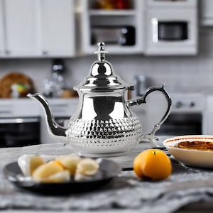 May include: A silver teapot with a textured surface and a rounded body. The teapot has a spout and a handle. The teapot is sitting on a table with a plate of fruit and a bowl.