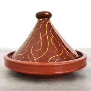 Op de afbeelding: Een terracotta tajine met een conische deksel, versierd met een decoratief ontwerp van draaiende lijnen in een gouden gele kleur.