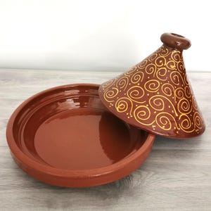Può includere: Una tagine in terracotta con un coperchio che presenta un motivo a spirale in vernice dorata. La tagine è una pentola da cucina tradizionale marocchina.