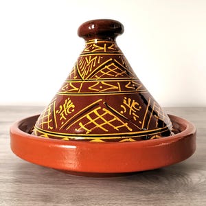 Peut inclure: Un tajine en céramique marron et orange. Le couvercle conique est orné de motifs géométriques jaunes. La base est un plat large et peu profond. Ce plat traditionnel marocain est conçu pour la cuisson lente.