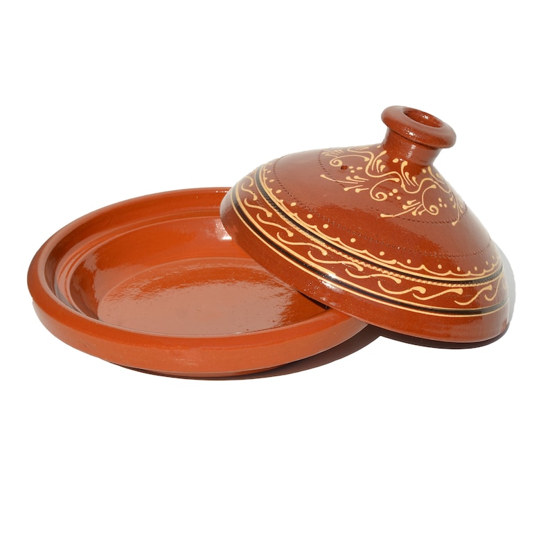 May include: A terracotta tagine with a lid featuring a decorative design of brown and gold swirls. The tagine is a traditional Moroccan cooking pot.