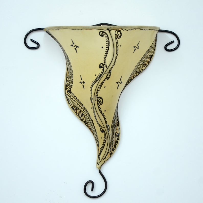 May include: A cream-colored wall sconce with a black metal frame and intricate black designs. The sconce is shaped like a teardrop and has a curved top.