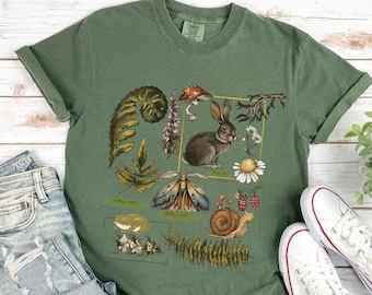 Könnte beinhalten: Ein grünes T-Shirt mit einem naturbezogenen Grafikdruck. Der Druck zeigt ein Kaninchen, einen Schmetterling, eine Schnecke, einen Pilz und verschiedene Blätter und Blumen.