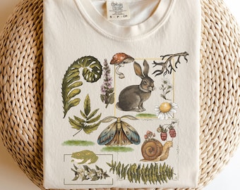 Könnte beinhalten: Ein cremefarbenes T-Shirt mit einer Vintage-Illustration eines Kaninchens, einer Schnecke, eines Schmetterlings, eines Frosches und verschiedener Pflanzen. Die Illustration ist von einem goldenen Rahmen umgeben.