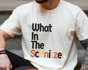 Może przedstawiać: Kremowa koszulka z napisem "What In The Schnitzel" w czarnych, pomarańczowych i żółtych literach. Słowo "Oktoberfest" jest wyświetlane na banerze u dołu. Koszulka jest noszona przez osobę.