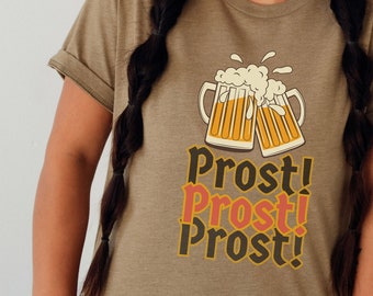Może przedstawiać: Oliwkowo-zielona koszulka z grafiką dwóch stukających się kufli piwa, z pienistym piwem i napisem "Prost!" w warstwowym wzorze. Koszulka ma podwinięte rękawy.