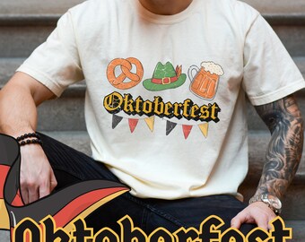 Może przedstawiać: Kremowa koszulka z motywem Oktoberfest. Grafika zawiera precla, zielony kapelusz i kufel piwa. Słowo "Oktoberfest" jest napisane w stylu vintage, z banerem flag poniżej.