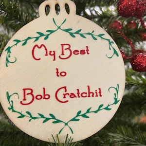 Puede incluir: Un adorno navideño de madera con un diseño de corona verde y el texto "My Best to Bob Cratchit" en rojo.