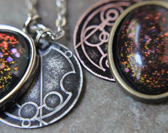 May include: A silver necklace with three pendants. The first pendant is a round, silver charm with a black and white design. The second pendant is a round, silver charm with a black and white design. The third pendant is a round, bronze charm with a black and orange design.