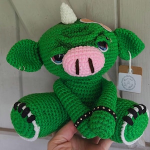 Puede incluir: Una criatura verde hecha a mano con ganchillo, con un cuerno blanco, un hocico rosa y ojos azules. Tiene detalles en blanco y negro en los pies y una pulsera negra con tachuelas. La criatura está sentada con las piernas cruzadas y tiene una etiqueta.