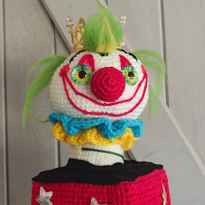 May include: A crocheted jack-in-the-box toy featuring a clown head with green hair, a gold crown, and a red nose. The clown wears a blue and yellow ruffled collar. The base is a red crocheted cube decorated with various colored glittery star appliques.